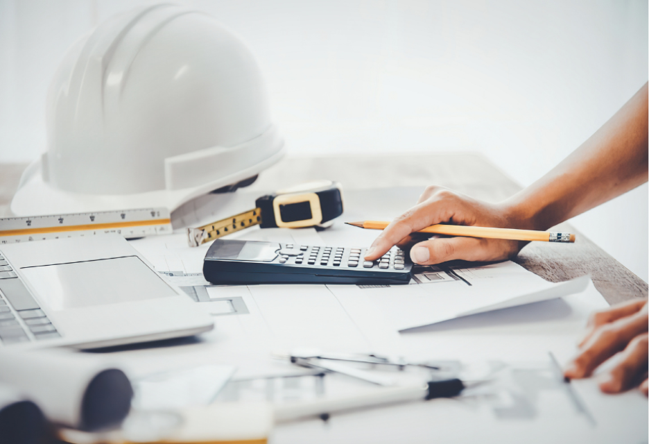 Construction professional using a calculator over architectural plans at a desk, with a hard hat, measuring tools, pencil, and laptop nearby.